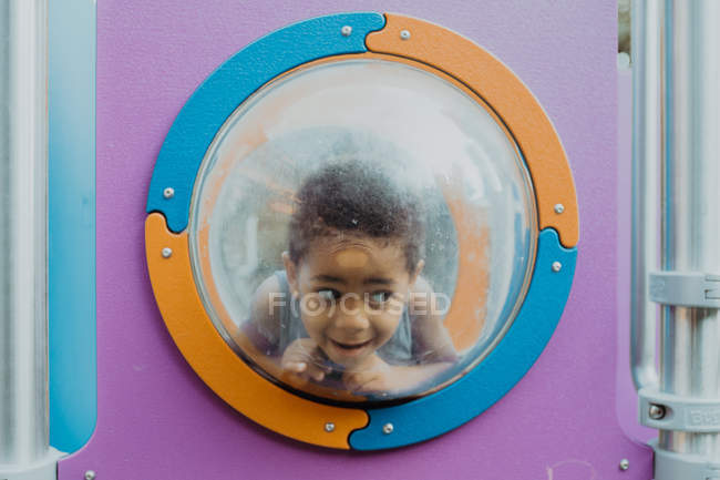 Piccolo ragazzo afroamericano guardando lontano attraverso oblò e facendo facce divertenti mentre gioca sul parco giochi — Foto stock