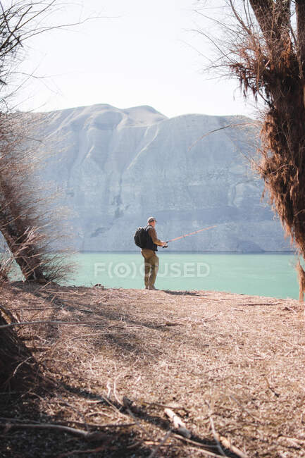 Back view of male with backpack standing on lake shore and fishing on sunny day in nature — Stock Photo
