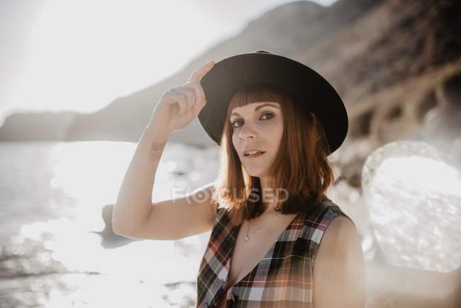 Attractive female with unbuttoned checkered shirt posing near sea water on rocky coast against mountains on sunny day in countryside — Stock Photo