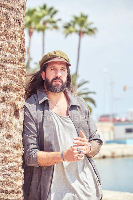 Stylish bearded man with long hair leaning on palm tree — Stock Photo