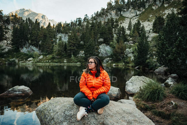 Schöne junge Frau in lässigem Outfit schaut weg, während sie auf einem Felsen in der Nähe des ruhigen Sees in der Natur sitzt — Stockfoto