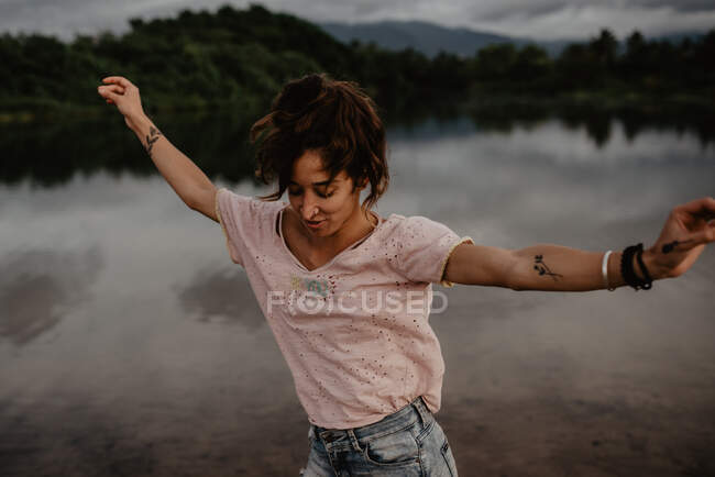 Вид сбоку взволнованной молодой женщины, улыбающейся и танцующей с закрытыми глазами и поднятыми руками у пруда со спокойной водой в сельской местности — стоковое фото