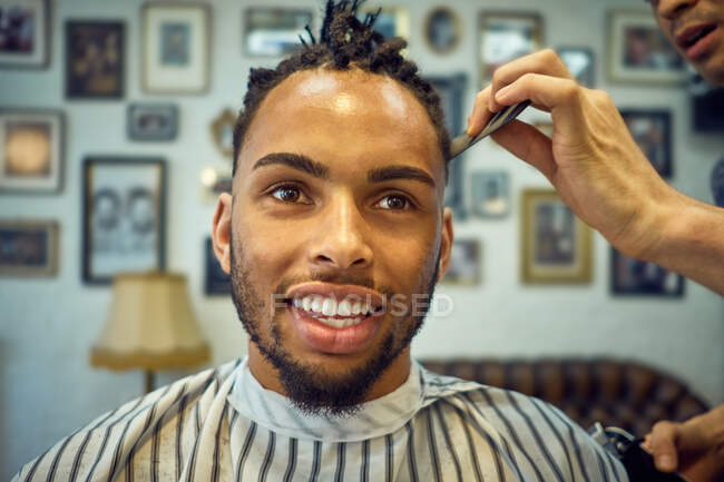 Vista da colheita de cabeleireiro anônimo fazendo um corte de cabelo moderno para um cliente afro-americano alegre — Fotografia de Stock