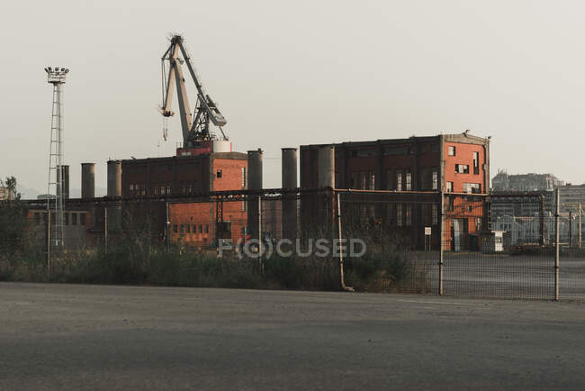 Edificios antiguos de fábrica de ladrillo rojo en funcionamiento, tuberías y grúas colocadas en el área industrial detrás de la valla - foto de stock
