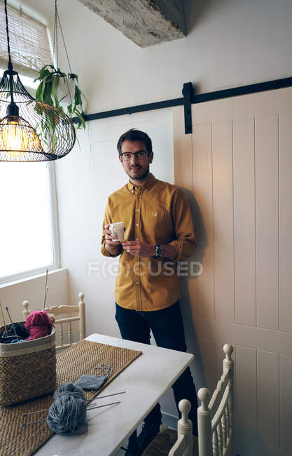 Adulto macho desfrutando de chá quente fresco e olhando para a câmera enquanto sentado à mesa perto de agulhas de tricô e fios — Fotografia de Stock