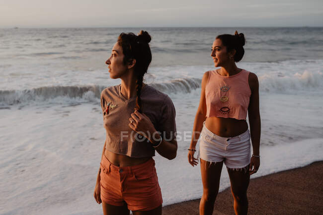 Zwei schlanke junge Frauen in kurzen Hosen und BHs schauen weg, während sie am sandigen Ufer vor wolkenverhangenem grauen Himmel bei Sonnenuntergang stehen — Stockfoto