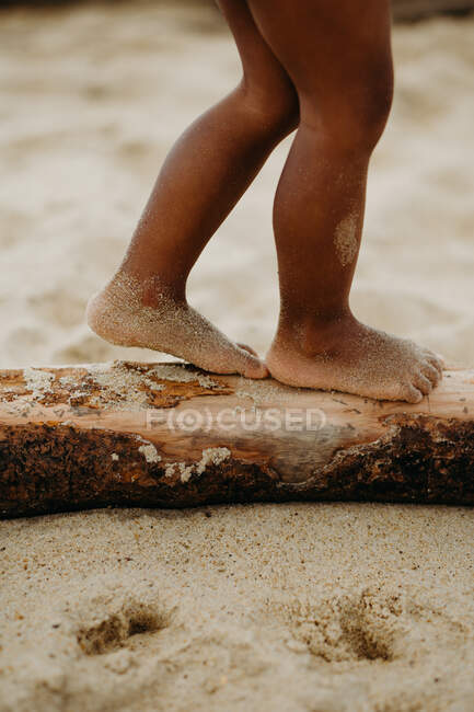 Pernas sujas de criança negra irreconhecível andando no log na praia de areia — Fotografia de Stock