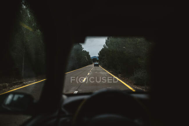 Вид на асфальт сільської дороги та зелені дерева лісу через лобове скло сучасного автомобіля під час подорожі на природі — стокове фото