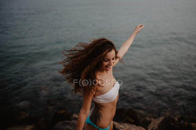 Vue latérale de jolie jeune femelle en maillot de bain souriant et dansant près de l'eau calme de la mer dans la nature — Photo de stock