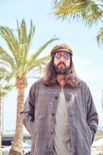 Stilvoller bärtiger Mann mit langen Haaren, der mit Sonnenbrille in der Nähe einer Palme auf der Straße spaziert — Stockfoto