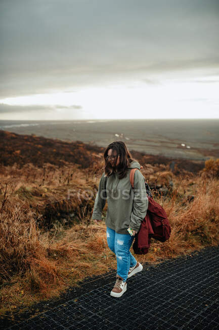 Young female hiker in comfort clothes with backpack walking in desert countryside against grey gloomy sky — Stock Photo