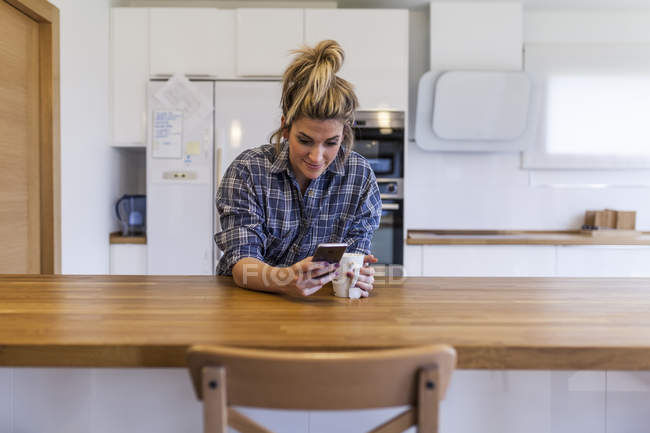 Bela e jovem mulher tomando café da manhã em casa e se divertindo e conversando com o smartphone — Fotografia de Stock