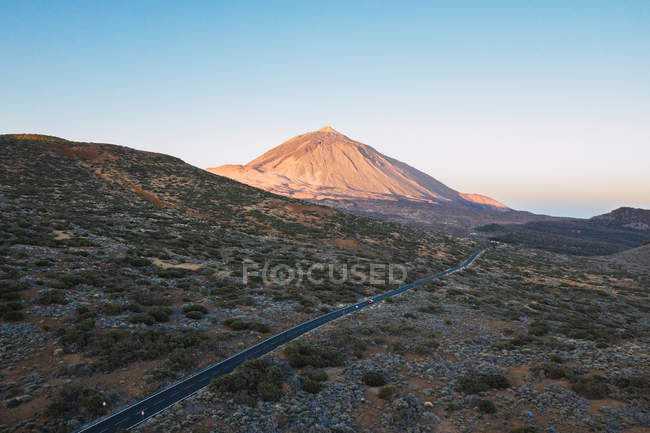 Vista panorâmica da montanha iluminada pico rochoso e estrada vazia na área do deserto contra céu crepúsculo claro — Fotografia de Stock