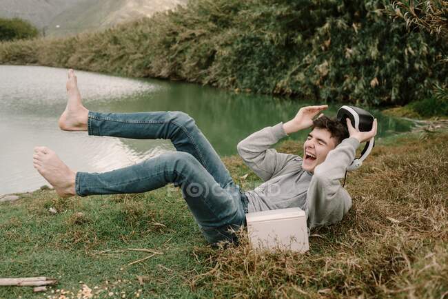 Adolescente joven con gafas de realidad virtual tumbado en la hierba al aire libre cerca de un lago con un libro y riendo en voz alta - foto de stock