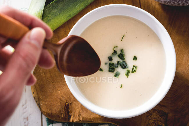 Shoot from above of crop hand with spoon and tasty Vichyssoise soup on wooden table with leek — Stock Photo
