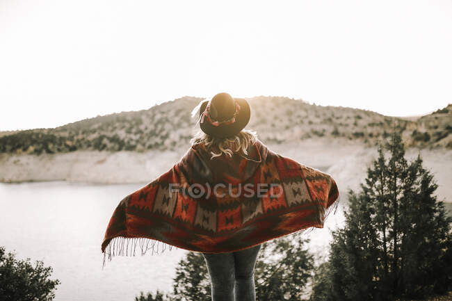 Back view of lady in hat and poncho looking at sunrise sky and mountains while standing on shore of tranquil lake — Stock Photo