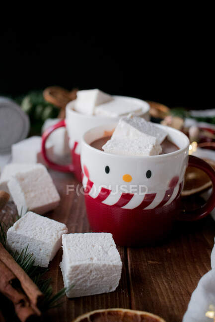 Ароматические палочки корицы и сушеные цитрусовые, помещенные на пилораме возле чашек вкусного горячего шоколада с мягкими зефирами и различными рождественскими украшениями — стоковое фото
