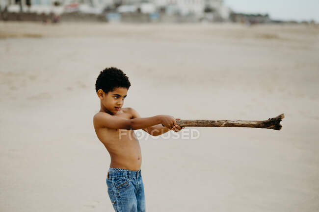 Divertido chico afroamericano con palo jugando en la orilla de arena cerca del mar - foto de stock