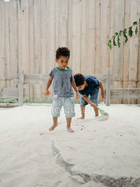 Dos adorables chicos afroamericanos con atuendos casuales jugando con arena seca mientras pasan tiempo en el patio en un día soleado - foto de stock
