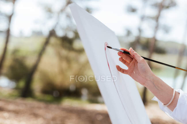 Mano de mujer anónima usando pincel para dibujar sobre lienzo sobre fondo borroso de la naturaleza - foto de stock