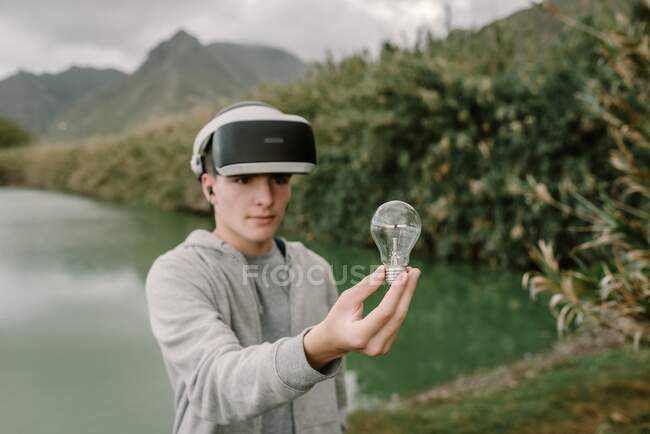 Joven adolescente jugando una simulación de realidad virtual con gafas vr de pie cerca de un lago sosteniendo una bombilla iinovación y concepto de energía - foto de stock