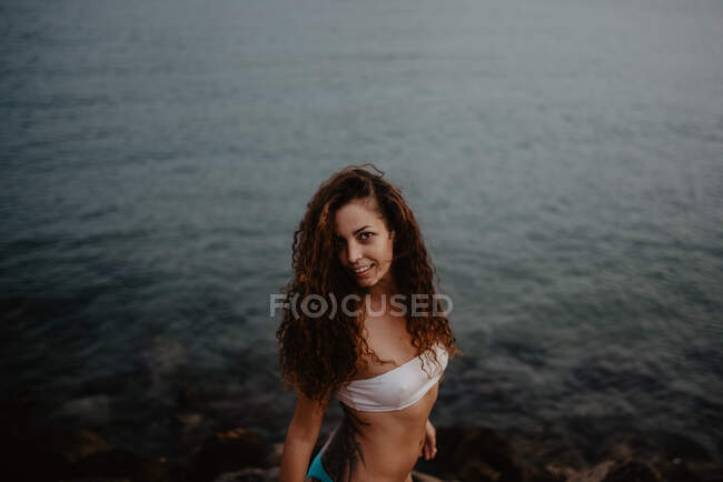Вид сбоку на симпатичную молодую женщину в купальнике, улыбающуюся и смотрящую в камеру, стоя у спокойной морской воды на природе — стоковое фото