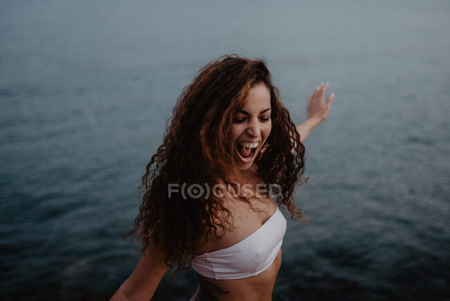 Вид сбоку на симпатичную молодую женщину в купальниках, улыбающуюся и танцующую рядом со спокойной морской водой на природе — стоковое фото