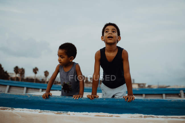 Два изумленных афроамериканских мальчика, опираясь на старое старое судно и отводя взгляд, проводили время на пляже вместе — стоковое фото