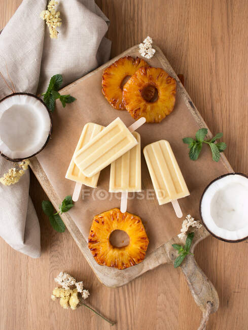 Scheiben frischer Ananas und Hälften reifer Kokosnüsse mit Minze um köstliches Eis an Bord in der Nähe von Serviette gegen Holztischplatte gelegt — Stockfoto