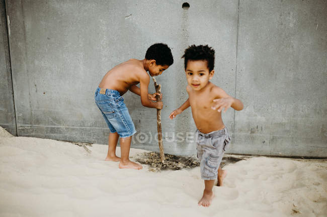 Dois irmãos afro-americanos com paus brincando juntos na costa arenosa perto do mar — Fotografia de Stock