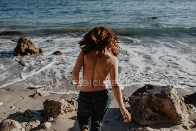 Back view of barefoot topless woman stepping towards foamy waves of stormy sea on sunny day in nature — Stock Photo