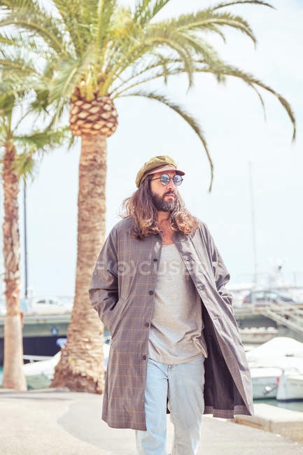 Elegante uomo barbuto con lunghi capelli che camminano sulla strada con occhiali da sole vicino alla palma — Foto stock