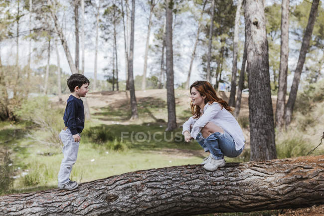 Side view of mother sitting on tree trunk and smiling while communicating with little boy on sunny day in countryside — Stock Photo