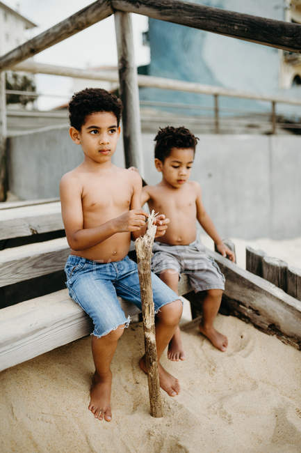 Два афроамериканских брата с палками сидят и играют вместе на песчаном берегу у моря — стоковое фото