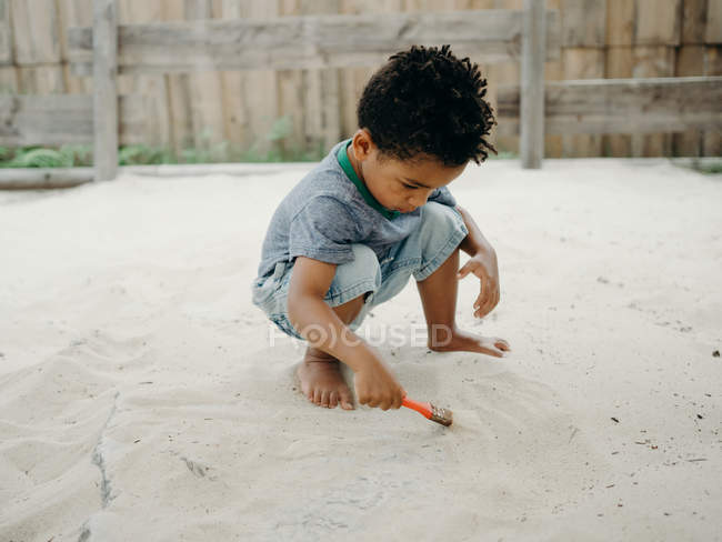 Adorable chico afroamericano en traje casual jugando con arena seca mientras pasa tiempo en el patio en día soleado - foto de stock