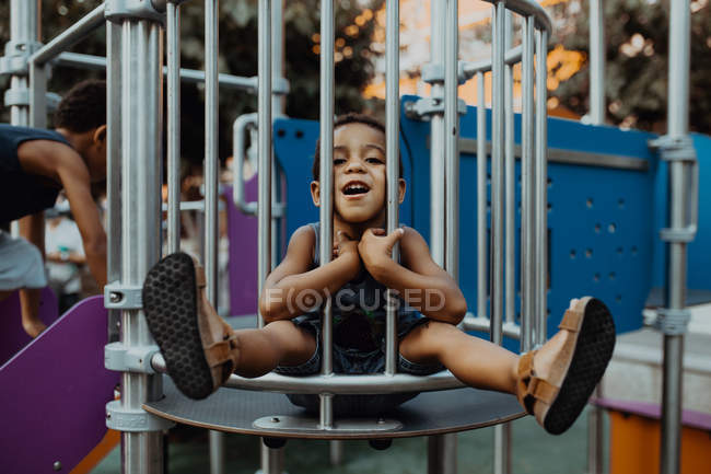 Afro-américain garçon avec drôle visage regardant caméra tout en étant assis derrière grille sur aire de jeux dans le parc — Photo de stock