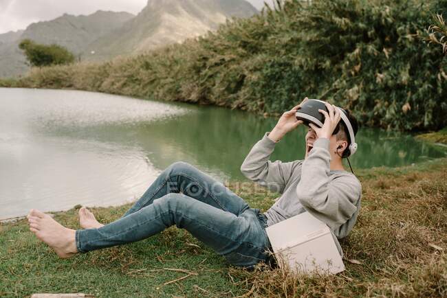 Young adolescent with virtual reality glasses laying on grass outdoors near a lake with a book — Stock Photo