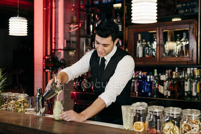 Jovem barman elegante trabalhando atrás de um balcão bar derramando bebida de agitador para um copo — Fotografia de Stock
