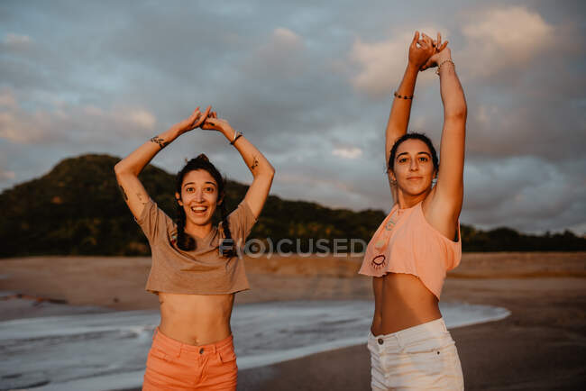 Zwei schlanke junge Frauen in kurzen Hosen und BHs, die lächelnd in die Kamera schauen, während sie die Arme am sandigen Ufer vor dem wolkenverhangenen grauen Himmel bei Sonnenuntergang ausstrecken — Stockfoto
