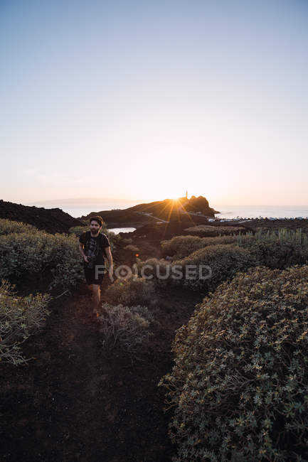 Homme voyageur avec caméra marchant sur le bord de mer désert lointain en Espagne contre ciel sans nuages et coucher de soleil lumineux — Photo de stock