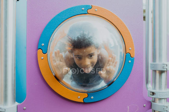 Little African American boy looking away through porthole and making funny faces while playing on playground — Stock Photo