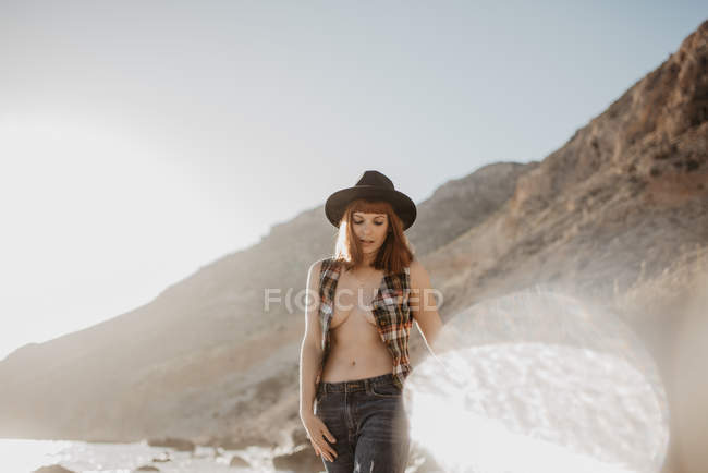 Mulher atraente com camisa quadriculada desabotoada andando perto da água do mar na costa rochosa contra montanhas no dia ensolarado no campo — Fotografia de Stock