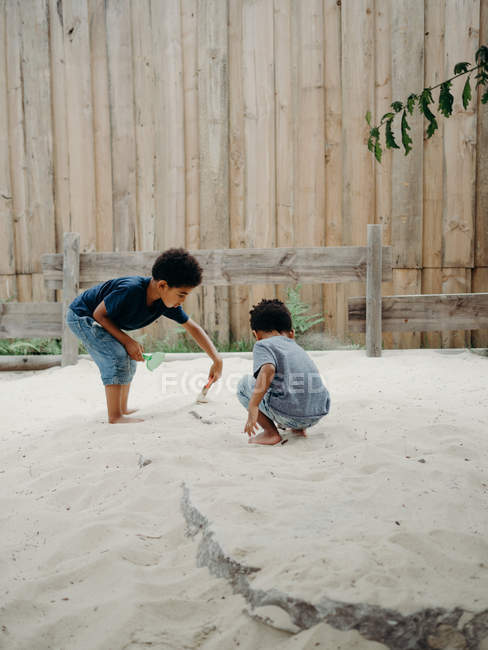 Два очаровательных афроамериканских мальчика в повседневной одежде играют с сухим песком, проводя время во дворе в солнечный день — стоковое фото