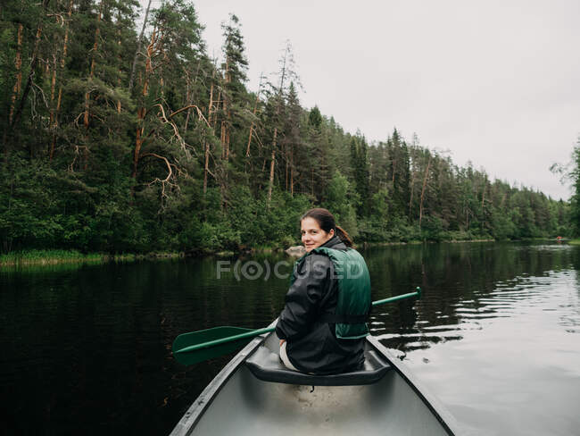 Joven mujer sonriente mirando hacia atrás mientras navega en el río bosque en Finlandia - foto de stock