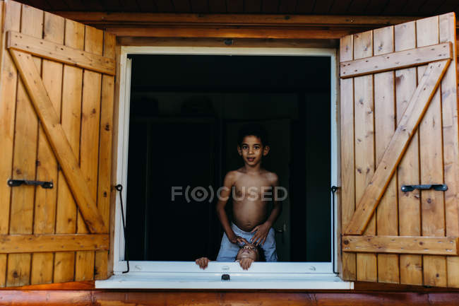 Dois irmãos afro-americanos sem camisa olhando para fora janela aberta de casa de madeira, enquanto se divertindo em casa juntos — Fotografia de Stock