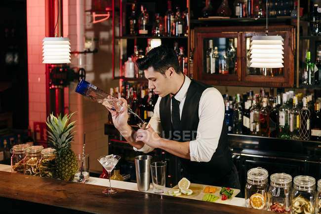 Jeune barman élégant travaillant derrière un comptoir de bar mélangeant boissons et fruits — Photo de stock