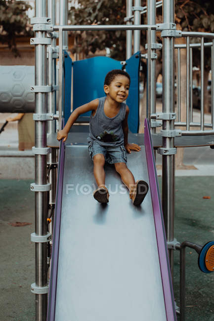 Adorabile bambino afroamericano in abito casual sorridente e seduto sullo scivolo sul parco giochi sulla strada della città — Foto stock