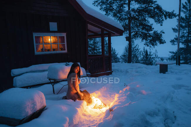 Vista lateral de mujer desnuda joven con tatuajes sentados en la nieve contra la casa rural mirando hoguera cerca - foto de stock