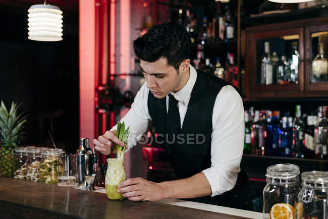 Jovem barman elegante trabalhando atrás de um balcão de bar preparando bebida em um copo — Fotografia de Stock