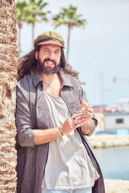 Stylish smiling bearded man with long hair leaning on palm tree — Stock Photo